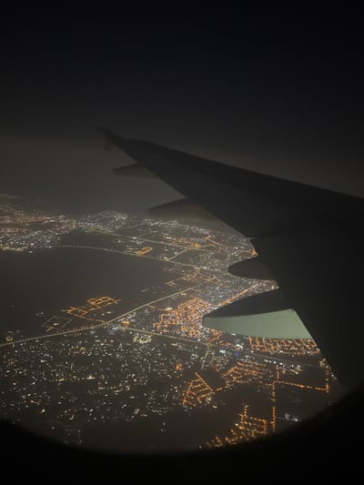 An airplane wing is visible through a dark, possibly fogged window. Below, a city sprawls with a dense network of illuminated streets, creating a vibrant display of orange and yellow lights. The city lights extend towards the edge of the window, partially obscured by darkness.