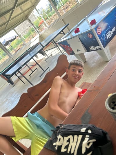 A young boy, shirtless, lies on a wooden picnic table, his head resting on his arm. He is wearing bright yellow shorts and a pair of light blue swim trunks pulled up over them. A red packet of snacks rests on his chest. Beside him, a black backpack with white lettering hangs from the table. In the background, a climbing wall with "Finding Nemo" imagery is visible, alongside windows offering a view of a sunny outdoor scene with trees and foliage. The image is taken from a slightly elevated angle, capturing the boy and the surrounding environment. The overall impression is one of a relaxed outdoor setting, possibly a park or a recreational area.