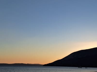 The image depicts a serene coastal scene at dusk, likely in Erdek, Turkey. The sky transitions from a clear, pale blue at the top to a soft gradient of orange and yellow near the horizon. In the foreground, a calm body of water, possibly the sea, is visible, with subtle ripples indicating gentle movement. Silhouetted against the colorful sky are the dark outlines of distant landmasses or islands, characterized by undulating, hilly terrain. On the water, a few small boats are present, adding a touch of human activity to the otherwise natural landscape. One boat, closer to the foreground and slightly right of center, appears to be a fishing vessel, while other smaller, lighter shapes suggest buoys or additional vessels further out. The overall impression is one of tranquility and the quiet beauty of a coastal sunset.