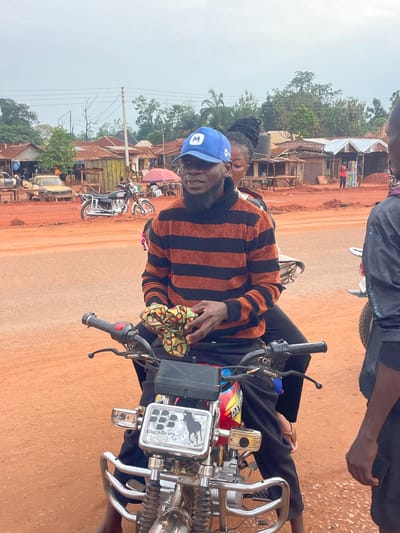 The image shows a man wearing a blue cap and a striped orange and black sweater, seated on a motorcycle in Otukpa, Nigeria. A woman is seated behind him, partially visible. The motorcycle features a rectangular aftermarket light fixture labeled "Blackberry POLO." The setting appears to be a dirt road with various buildings in the background, suggesting a market or commercial area. Other motorcycles are parked nearby. A person wearing a black shirt is standing adjacent to the motorcycle. The ground is a reddish-brown color.