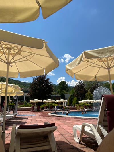 The image depicts a sunny day at an outdoor swimming pool, likely within a resort or hotel in Zverino, Bulgaria, given the context. In the foreground, two large, cream-colored umbrellas dominate the view, casting shadows on the red-tiled patio. Several white lounge chairs are arranged on the patio, some empty and some occupied by people.

The swimming pool, filled with blue water, is visible in the mid-ground, with several individuals swimming and playing. A decorative fountain with a spherical, metallic structure is located at the far end of the pool.

In the background, more umbrellas and lounge chairs are set up. Beyond the pool area, there is a landscaped garden with various trees, including dark-leaved trees and evergreens. A building with a Coca-Cola sign is visible on the left side of the frame. Further in the distance, rolling hills covered with trees and some scattered buildings suggest the surrounding mountainous terrain typical of the region. The sky is a clear blue with scattered white clouds.