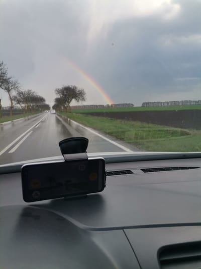 The image captures a first-person view from inside a car, looking out over a wet road and rural landscape during an overcast day. A vibrant rainbow arcs across the sky on the right side of the frame.
In the foreground, the dark grey dashboard of the car is prominent, featuring a textured surface and air vents. A black smartphone mount holds a smartphone horizontally on the dashboard. The phone's screen displays several user interface elements, including circular yellow icons (one with a possible play/record symbol, another with a warning triangle) and a small white square icon in the top right, but no clearly legible text.
The main subject of the scene is a two-lane asphalt road, which appears wet and reflective from recent rain. Double white lines mark the center of the road. On both sides, lines of bare-branched trees, possibly forming an avenue, stretch into the distance. Several cars with headlights on are visible in the distance, suggesting ongoing traffic.
Beyond the road, to the right, are large agricultural fields with patches of green vegetation and dark brown earth. Another distant line of trees marks the horizon. The sky is predominantly grey and cloudy, typical of post-rain weather, but lightens where the rainbow is most prominent. The rainbow is distinct, its colors clear against the muted sky, starting from the mid-right and extending towards the upper right of the image.
The scene depicts driving through a rural area, consistent with the location context of Stefanovo, Bulgaria. The time of day is daytime, characterized by overcast skies and the aftermath of rain, creating the conditions for the rainbow's appearance.