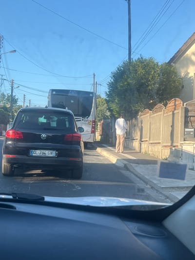 The image captures a street scene in Gagny, France, likely on a sunny day given the clear blue sky. In the foreground, a black Volkswagen Tiguan is parked, its rear visible, displaying a French license plate: CL-794-GN. Behind it, a white bus, possibly a public transport vehicle, is also stopped. To the right of the bus, a person dressed in a white long-sleeved shirt and beige pants stands on a sidewalk next to a fence. The fence appears to be made of concrete and wood, with arched wooden panels at the top. The street is paved, and a sidewalk runs along the right side of the image. Overhead, power lines and a streetlight are visible against the sky. The overall impression is that of a quiet suburban street during daylight hours.