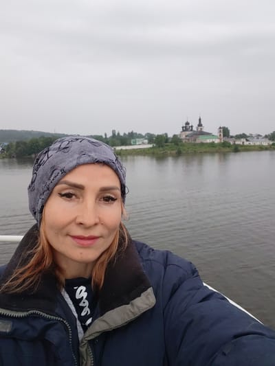 A woman wearing a gray patterned beanie, a dark blue jacket, and a dark t-shirt is pictured in a selfie on a body of water. The woman has light-colored hair, prominent eye makeup, and is smiling faintly. In the background, across the water, is a complex of buildings with multiple domes, consistent with the Goritsy Monastery in Goritsy, Russia. The sky is overcast and the water appears calm. The perspective suggests the photo was taken from a boat or a similar watercraft.