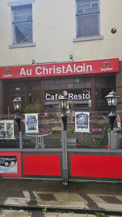 This media file captures the exterior of a cafe or restaurant named "Au ChristAlain" in Thuin, Belgium. The scene is viewed from across a wet pavement, suggesting recent rain or damp conditions.
The establishment features a prominent red banner sign along its facade with "Au ChristAlain" written in white lettering, accompanied by the Jupiler beer logo. Below this, the entrance area consists of large glass windows and a dark-framed door. Inside the windows, another sign reads "Café Resto" in white and purple. Various posters are displayed on the glass, one with the text "Passez à l'arrière" (Go to the back) and "Terrasse à l'arrière" (Terrace at the back), indicating additional seating. Other posters show images of the interior or dishes offered, and one clearly mentions "Thuin" as part of an address or contact information.
Outdoor seating is visible through the glass, including small tables and chairs, some green and white. Two decorative lampposts are positioned outside the entrance. The building itself is light-colored, possibly painted brick or stucco, with two blue-framed windows on the upper floor. The pavement directly in front of the establishment features red rectangular sections integrated into the ground, possibly part of an outdoor dining area or a decorative element. Small weeds are growing in cracks in the concrete pavement. The time of day appears to be daylight, but without direct sunlight.
Visible text includes:
- "Au ChristAlain"
- "Jupiler" (logo and text)
- "Café Resto"
- "Passez à l'arrière"
- "Terrasse à l'arrière"
- "Rauration THUILLIES"
- "Tel: 071 591 390"
- "Salle ouvert jusque 16h" (Room open until 4 PM)
- "Réunions et arrivées" (Meetings and arrivals)
- "La Lutèce" (on a poster)
- "10h" (possibly an opening time)