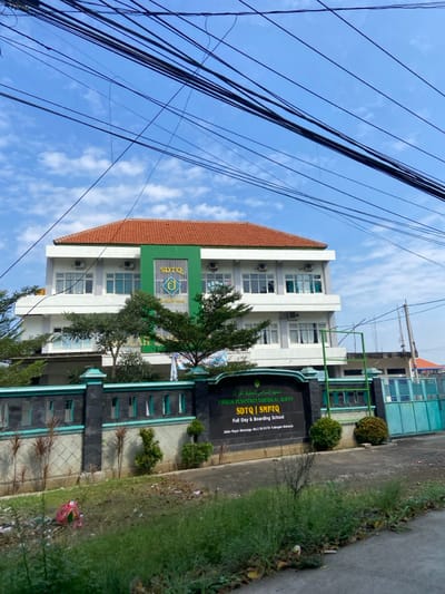 The image shows a two-story school building, identified as "SDTQ | SMPTQ Full Day & Boarding School", located at Jalan Raya Kemanggisan No.21 in Tulangan, Sidoarjo, Indonesia, with coordinates of -7.487 latitude and 112.654 longitude. The building is white with a red tile roof and green accents. It is surrounded by a black and green fence. A small, green tree grows in front of the building. The building's facade is visible, with windows and an entrance. The school's sign is prominent and displays the school's name, address, and information about being a full-day and boarding school. There are many power lines running across the sky, and the sky is a clear blue with a few white clouds.