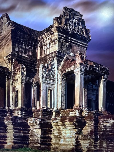 The image depicts the exterior of a large, ancient stone temple with intricate carvings and tiered structures. The architecture suggests Southeast Asian influence, possibly Khmer. The stone appears weathered, with some sections dark and others illuminated by dramatic lighting, casting deep shadows. The sky above is a deep purple with streaks of cloud, indicating dusk or dawn. The foreground shows hints of vegetation. The location provided, Urbanització Vescomtat De Cabanyes, Spain, is inconsistent with the architectural style of the temple, suggesting a potential misidentification of the location. There are no people or animals visible in the image.