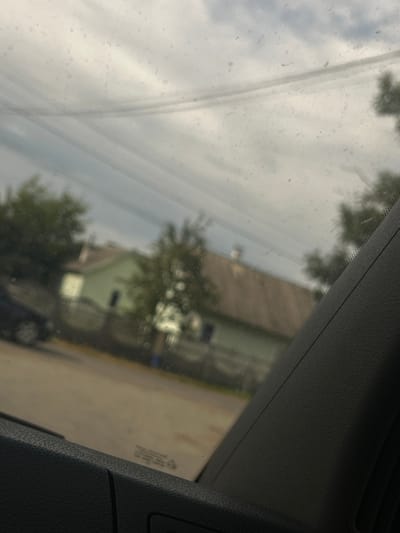 The image provides a view from inside a vehicle looking out through a dusty window. The scene depicts a residential area with a pale green house with a grey roof in the background, partially obscured by trees. A dark-colored car is visible on the left, and a section of a black interior panel of the vehicle occupies the foreground on the right. The sky is overcast with grey clouds. The location is identified as Yizhivtsi, Ukraine.