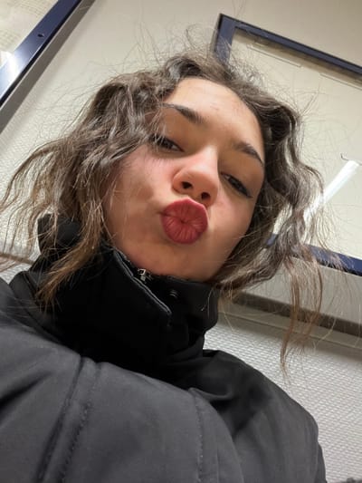 A young woman with wavy brown hair puckers her lips in a selfie. She wears a black jacket zipped up to her neck. The image is tilted, with the woman's head angled towards the lower left corner. In the background, a wall with a blue-framed window is visible. The lighting is diffused.