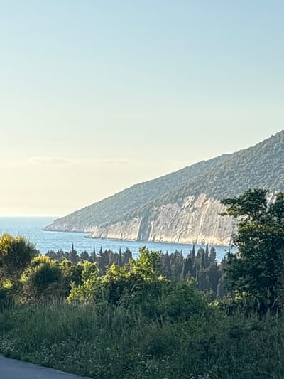 The image depicts a coastal scene in Bratica - Braticë, Montenegro. A large, rocky cliff dominates the frame, descending towards the Adriatic Sea. Vegetation, including trees and shrubs, grows along the cliff's edge and the right side of the image. The sky is pale and bright.