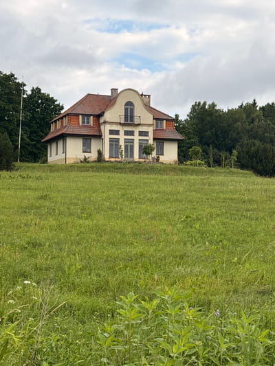 The image shows a large, light-colored house with a red tiled roof, situated on a grassy hill. The house has multiple windows, a balcony, and appears to be surrounded by trees. The foreground of the image is dominated by a field of tall green grass with some wildflowers. The sky is overcast with scattered clouds. The location is Aukštieji Rusokai, Lithuania.
