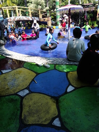 The image depicts a lively scene at a water park, likely located in Tegalwangi, Indonesia, given the lush tropical foliage visible in the background and the style of the park. The central focus is a swimming pool filled with clear blue water, teeming with children and a few adults. Several children are actively engaged in water play, some using inflatable inner tubes. One child, wearing a blue swimsuit, is partially submerged in an inner tube. Another child in a pink top is being held by an adult. A child in a red shirt is also visible in the water. The surrounding area of the pool features a colorful, mosaic-like concrete floor with large, irregularly shaped sections painted in vibrant yellow, green, and blue. The upper portion of the image reveals a wooden bridge structure, likely part of the park's attractions, with tropical trees and foliage behind it. White sandals are placed on the edge of the pool, near the colorful flooring. The overall atmosphere suggests a recreational and family-friendly environment.