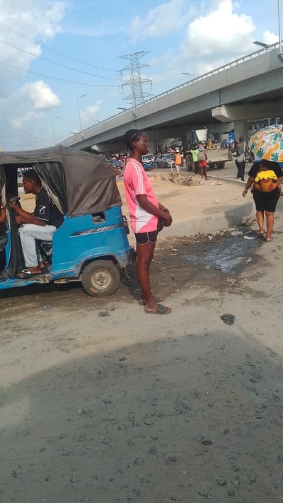 The image depicts an outdoor scene in Effurun, Nigeria. In the foreground, a woman wearing a pink shirt and black shorts stands next to a blue "Keke Napep" (tricycle) with a passenger inside. A bridge and an electrical tower are visible in the background, along with other pedestrians, vehicles, and construction equipment. The ground is dusty and appears to be under construction. The lighting suggests it is daytime under a partly cloudy sky.