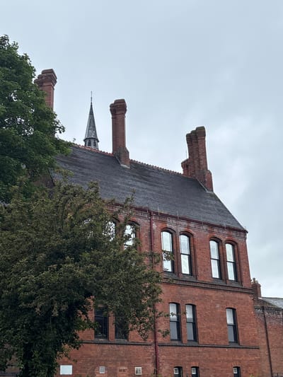 The image displays a large, distinctive brick building against an overcast sky, partially obscured by green trees. The scene is viewed from an upward angle, with the main building dominating the upper right portion, and a church spire visible in the background.
The primary subject is a multi-story building constructed from reddish-brown brick. Its roofline features several prominent, decorative brick structures that extend outwards, possibly elaborate chimneys or architectural embellishments. The roof itself is dark and appears textured, likely made of slate or tiles, sloping downwards. The facade exhibits various window styles; on the left section of the building, there are stacks of three windows, each with an arched top and flat bottom, set within a larger arched brick recess. Further to the right, other windows are more rectangular, some with slightly rounded corners, and smaller square or rectangular vents are scattered across the brickwork.
Dense green foliage from numerous trees fills the foreground and mid-ground, obscuring the lower parts of the building. Peeking above the trees and behind the main building's roof, a dark, slender church spire with architectural details is visible, suggesting another structure nearby.
The sky is a uniform light grey, indicating an overcast day. No human activity or visible text is present in the image. The architectural style of the brick building and the presence of a church spire are consistent with historic buildings found in the United Kingdom, aligning with the given location context of Knutsford.