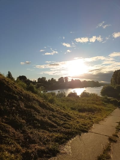 The image depicts a sunlit river landscape under a blue sky with scattered clouds, possibly during the late afternoon or early evening. The sun is positioned high in the sky, casting a strong glare on the water and illuminating the scene. A grassy, mown embankment occupies the foreground on the left, sloping down towards a paved pathway that runs along the right side of the image. Beyond the embankment and path, a river or large body of water is visible, bordered by a line of trees on the opposite bank. The trees are dense and lush, suggesting a natural or semi-natural environment. The lighting suggests a warm, clear day. The location is identified as Buy, Russia.