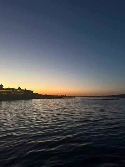 A photograph captures a tranquil body of water at dusk, with a gradient of colors in the sky ranging from deep blue at the top to a warm orange and yellow near the horizon. On the left side of the image, a long, low building with illuminated windows sits on the shore, silhouetted against the bright sky. A few scattered birds are visible in the sky. In the distance, across the water, a series of lights line a pier extending into the sea. The water itself is dark and rippled, reflecting the subdued colors of the sky. The scene appears to be located in Kazashko, Bulgaria.