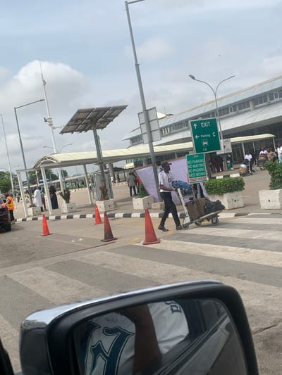The image, taken from inside a car, depicts a busy outdoor area at what appears to be an airport in Gwaska, Nigeria. The scene is set during the daytime, under a bright sky with scattered clouds.
In the foreground, a car's side mirror is visible on the right, reflecting part of the car's interior. A pedestrian crosswalk with alternating light and dark grey stripes stretches across the asphalt road. Several orange traffic cones are placed along the edge of the road, delineating a path or restricted area.
The central subject is a man, dressed in a light-colored collared shirt and dark trousers, actively pushing a metal luggage cart laden with bags. He walks along the crosswalk, seemingly moving towards or away from a building. Behind him and further in the background, various people are visible, some appearing to wait or walk near a building facade, which suggests an airport terminal. Concrete planters with greenery and large concrete blocks are also present.
Overhead, multiple street lights and poles with mounted equipment, possibly solar panels, are visible against the sky. A prominent green road sign indicates "↑ EXIT" and "← Parking C" with a "Car" icon. Below it, another green sign displays regulations: "FAAN", "NO PARKING", "NO WAITING", "NO PHONING", "KEEP MOVING", and warns of a "PENALTY = #25,000", confirming the Nigerian context. The overall activity suggests a dynamic environment typical of an airport drop-off or pick-up zone.