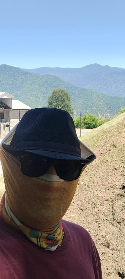 A person wears a dark blue fedora hat, dark sunglasses, and a yellow patterned face covering. The person is wearing a maroon sweatshirt. The background shows a lush green mountainous landscape under a clear blue sky. To the left, a portion of a building with a white roof and gray walls is visible. The ground in front of the person is brown and appears to be soil. The scene is set outdoors in a rural or mountainous area. The location of Milisi, Georgia is not explicitly indicated by any visual cues in the image.