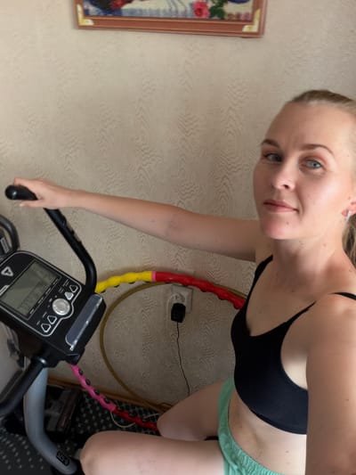 A woman with blonde hair pulled back, wearing a black sports bra and mint green shorts, sits on an exercise bike indoors. She is looking towards the camera with a slight smile. Her right arm is extended, resting on the bike's handlebars. The exercise bike's console displays various metrics. Behind the bike, a hula hoop with yellow, red, and pink sections is visible on the floor, along with an electrical outlet and plug. The wall behind the subjects has a textured wallpaper pattern. A framed picture hangs on the wall in the upper portion of the image. The overall scene appears to be a home environment within the city of Kul'dur, Russia.