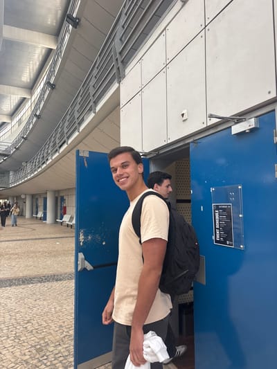 The image displays an outdoor or semi-outdoor area of the Nova School of Business and Economics (Nova SBE) in Carcavelos, adjacent to Porto Salvo, Portugal. A young adult male with dark hair and light skin stands prominently in the foreground, smiling directly at the camera. He wears a light beige short-sleeved t-shirt, dark grey trousers, and a black backpack, while holding a crumpled white garment in his left hand.

He is positioned at the threshold of a pair of partially open blue doors. Behind him, inside the doorway, a second young adult male with dark hair is partially visible, wearing a dark shirt and backpack, looking towards his right (viewer's left).

The architectural environment consists of a modern, multi-level building featuring a curved facade with grey metal railings and pillars supporting overhead structures. The ground is paved with a light-colored cobblestone pattern. Along the walkway to the left, several white benches are visible, and multiple individuals are walking in the distance. Above the blue doors, a white panel wall incorporates door closers and a small red indicator light. To the right of the doors, a clear acrylic display holder contains a black and white "EVENT SCHEDULE" notice for "SATURDAY 16-18," featuring the Nova SBE logo.