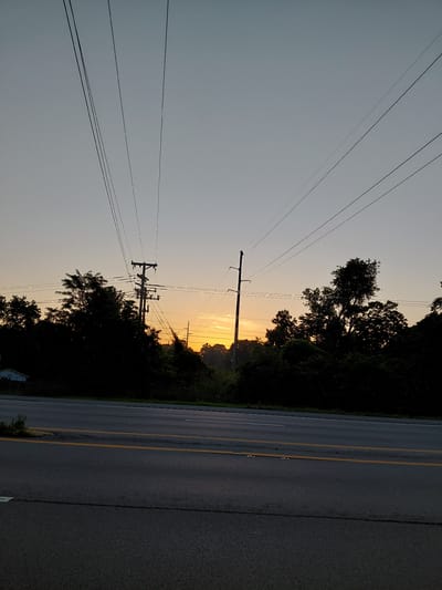 The image captures a view of a road and power lines against a dusky sky, likely at sunrise or sunset. The sky is a pale gradient of grey and orange, with the sun's glow visible near the horizon, partially obscured by silhouetted trees and utility poles. Multiple power lines stretch across the frame, originating from two prominent utility poles. One pole is situated on the right side of the image, with the other slightly to the left, closer to the center. Dense, dark foliage and trees form a thick line along the horizon, obscuring the ground behind them. In the far left background, a small, light-colored building, possibly a shed or small house, is partially visible through the trees. The foreground shows an asphalt road with yellow and white lane markings. The road appears to be a multi-lane highway. The scene is devoid of people or any discernible activity. The overall impression is a quiet, somewhat desolate roadside landscape during twilight. The location is not specified in the image itself and therefore cannot be identified as Frankfort, United States of America.