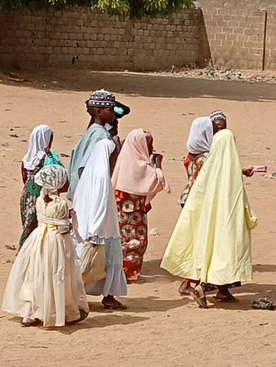 The image displays a group of approximately seven to eight individuals, primarily children and young people, gathered or walking on a sandy, unpaved path in Fanisau, Nigeria. The bright lighting suggests it is midday or early afternoon, with clear shadows cast on the light brown earth.
The subjects are dressed in traditional attire. On the far left, a young girl wears a long, light cream-colored dress with a gathered waist and a head covering, facing away from the viewer. Next to her, another individual is in a white head covering and a light blue garment. A young boy in the center sports a dark, intricately patterned Kufi cap and a light blue top. Other figures, mostly girls or women, are seen in various colored hijabs and flowing dresses: one in a pale pink hijab over a patterned red dress, another in a white hijab and a light blue skirt, and a partially obscured person in a green patterned outfit. On the far right, a figure in a bright yellow, full-body covering holds a small, light-colored rectangular object.
The background features a low, weathered brick wall made of light-colored blocks, likely part of a building or compound, with sparse green foliage from trees or bushes visible above it. The dusty ground and traditional clothing are consistent with the location context. There is no visible text in the image. The scene captures a casual moment of daily life in an outdoor community setting.