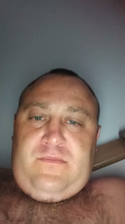The image is a close-up, low-angle selfie of a man's face, taken from the chest up. The man has fair skin, short dark hair, and blue eyes. His head is tilted slightly upwards, and he appears to be looking directly at the camera. He is shirtless, and his chest and neck are covered in dark hair. The background is a plain, light gray wall. There is a hint of a wooden object to the right of the man's head. The lighting suggests the photo was taken indoors, possibly in the evening or in a dimly lit room. There is no identifiable location in Žilina, Slovakia within the image.