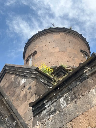 The image shows the dome of the Saint Sion Church, located in Artashavan, Armenia. The dome is constructed of light brown stone blocks and is round in shape. There are small plants growing on the dome and yellow flowers are visible on the adjacent stone structure. The sky is blue with scattered white clouds. A portion of the stone structure with dark grey accents is visible in the foreground.
