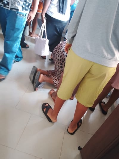 The image shows a group of people in a tiled room in Ogboko, Nigeria. A person wearing a leopard print dress is kneeling on the floor, with sandals stacked under their knees. Several other people are standing around them, some wearing sandals. There's a person in blue jeans walking past, and others in shorts and dresses. A white tote bag is visible among the standing people. The floor is tiled with light-colored square tiles.