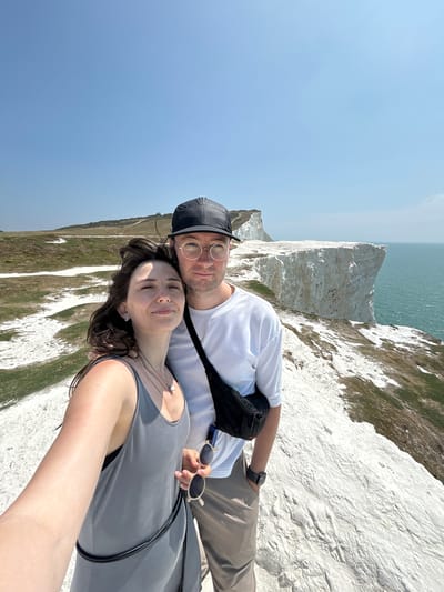 A couple is captured in a selfie on a cliff edge overlooking the ocean in Seaford, United Kingdom. The woman, on the left, is wearing a gray tank top and necklaces. The man, on the right, is wearing a white t-shirt, a black fanny pack, and a black baseball cap. They are both smiling at the camera. The background shows a vast expanse of white cliffs and the turquoise ocean. The sky is clear and blue. The ground is covered in chalky white material and sparse green grass. In the distance, other chalk cliffs and a pathway are visible. The sea is calm.