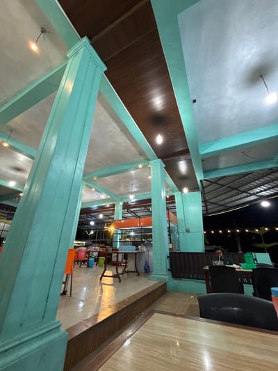 This media file captures an interior view of what appears to be a restaurant or cafe in Grong-Grong, Indonesia, at night. The scene is dominated by large, light blue painted wooden beams and columns, creating an open, airy architectural structure. The ceiling is a light color, possibly concrete or plaster, with visible lighting fixtures casting a warm glow.

The main subjects are the architectural elements and the ambient lighting. There is one person visible in the background, a man sitting at a table, appearing to drink from a straw. He is wearing headphones and a dark shirt. The setting includes tables and chairs, with some chairs being brightly colored (orange, green, blue) in the mid-ground. The floor is tiled with a wood-grain pattern. In the far background, through a glass partition, more tables and lighting are visible, suggesting a larger dining area.

Notable details include the artificial lighting, indicating it is nighttime. The overall mood is relaxed and inviting, typical of an evening dining establishment. The specific location cues are limited to the mentioned architectural style and the Indonesian context provided.

No discernible text is clearly visible in the image, though a sign that might contain text is partially visible in the far background.
