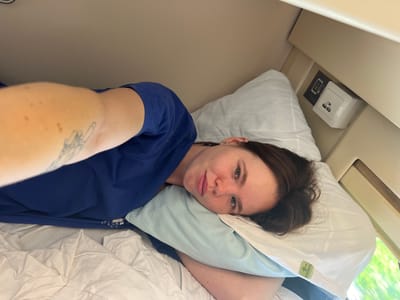 The image shows a person lying on a bed in a small room. The person is wearing a blue t-shirt and has a tattoo on their arm. There are two pillows on the bed, one white and one light blue. In the background, there is a charging station with USB ports. The room appears to be a sleeping cabin, possibly on a train or in a compact living space. The overall lighting suggests it is daytime, with natural light coming from a window on the right side of the image. The scene is static and depicts a moment of rest. The location is Urman, Russia.