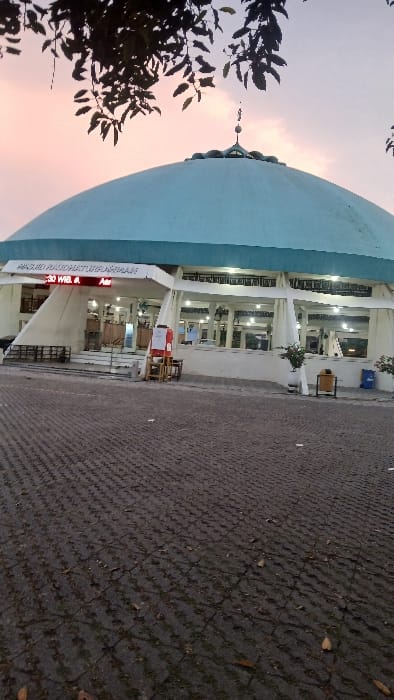 The image displays the exterior of Masjid Raudhaturrahmah in Pasar Paloh, Indonesia, during the evening. A large, light-blue dome with a finial crowns the white-walled mosque, which features multiple illuminated arched windows and openings. In the foreground, a dark gray interlocking paver surface shows sparse grass and scattered dry leaves. The main entrance, accessed via a short staircase, is marked by an overhead sign reading "MASJID RAUDHATURRAHMAH". An electronic display below the sign shows "19:30 WIB. A" and "Asr". To the left of the entrance, a red stand with a white sign and a small wooden stool are positioned near dark-colored benches. To the right, potted plants are visible alongside yellow and blue trash receptacles. Silhouetted tree foliage partially obscures the upper dome against a sky displaying gradients of pink, orange, and light blue. No individuals are discernible in the scene.