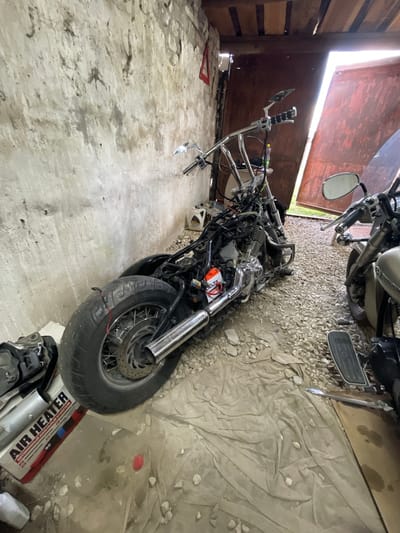 The image displays the interior of a garage or workshop in Znamensk, Russia, characterized by wooden plank walls and a mixed concrete and gravel floor. Bright daylight enters from the top left through an open, reddish-brown door, illuminating the scene.
The primary subject is a partially disassembled motorcycle lying on its side in the center. Its engine, chrome exhaust pipes, and intricate wiring are exposed, indicating ongoing maintenance or restoration. An orange box labeled "DELTA" is visible near the engine, and its front wheel is positioned towards the bottom right of the frame. In the upper right corner, parts of a second, more complete motorcycle are visible, including its handlebars, mirrors, and fuel tank.
The floor is a combination of rough concrete, sections covered with loose light-colored gravel, and a crumpled beige fabric or tarp. Various objects are scattered across the floor, such as a red triangular item on the concrete in the bottom left and a white container near the rear of the disassembled motorcycle. A cardboard box in the bottom right corner prominently features the English text "AIR HEATER" and Cyrillic text, "АВТОНОМНЫЙ ОТОПИТЕЛЬ," meaning "Autonomous Heater."
The strong natural light suggests it is daytime. The setting evokes a functional workspace dedicated to vehicle repair or modification. No people are present in the image.