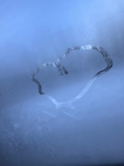 A heart-shaped outline is visible on a frosted glass surface in Ershovka, Russia. The heart is formed by a cleared or melted path through a layer of ice or condensation, creating a transparent channel against the opaque, muted blue-gray frosted background. The texture within the heart's outline shows slight variations, with some segments appearing as melted water droplets. The overall lighting is diffused. No individuals or other objects are present in the scene.