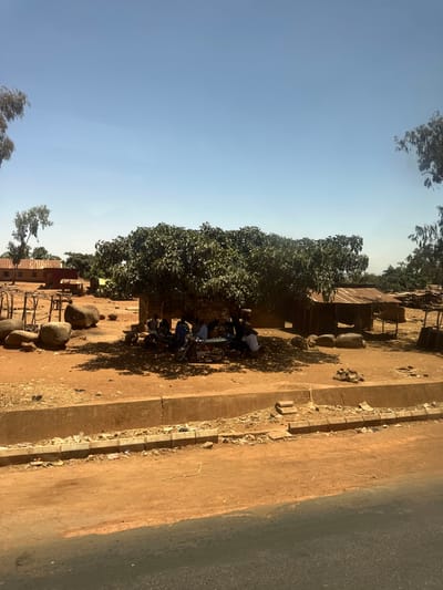 The image portrays a daytime outdoor scene in Hoss, Nigeria, under a clear, bright blue sky. The foreground shows a partial asphalt road, bordered by a concrete curb and an open drainage ditch containing assorted debris. The ground beyond the curb consists of reddish-brown, dry earth. A large, leafy green tree, centrally located, casts a dense shadow over the immediate area beneath it.
Under the tree's shade, approximately five to seven individuals are gathered, some seated, some standing, appearing to interact. Two motorcycles are parked among them. A small, light-colored table is visible in the center of the group. A faded inscription, possibly "MUMBA SAMA," is present on a low wall behind the gathering. To the left of the tree, several large, rounded boulders are scattered on the ground, along with a wooden, scaffolding-like structure.
In the mid-ground and background, multiple single-story buildings with reddish-brown corrugated metal roofs are visible, interspersed with sparse vegetation. To the right of the main tree, a smaller, open-sided structure with a corrugated metal roof stands on the reddish-brown earth. The scene is illuminated by strong, direct sunlight, evidenced by distinct shadows.