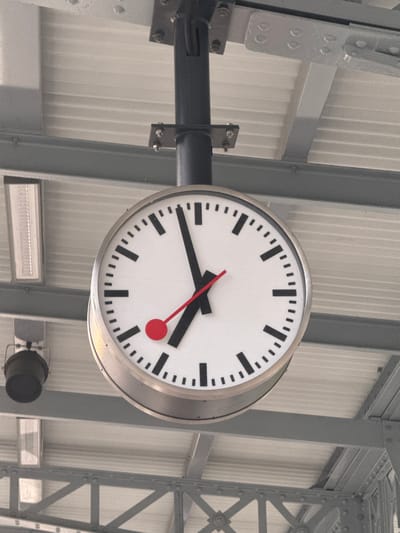 The image shows a close-up of a large, white, circular clock with black hour and minute markers. The clock has two black hands and a red second hand. The hour hand is pointing at approximately 10, and the minute hand is pointing at approximately 5. The clock is suspended from a dark pole, which is attached to a metal structure above, possibly part of a train station or public building. The background consists of a white, corrugated ceiling and grey metal beams. To the left of the clock, there is a black cylindrical object, possibly a speaker or light fixture, and a rectangular light fixture. The overall scene appears to be an outdoor or semi-outdoor public space. This clock is located at the train station in Sonceboz-Sombeval, Switzerland.