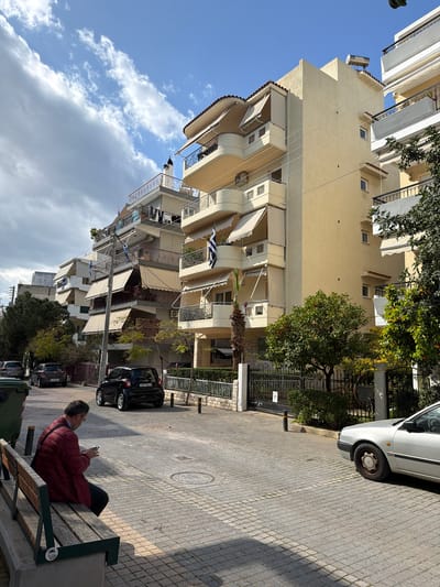 This image captures a sunny street scene in the Municipal Unit of Elliniko, Greece, featuring multi-story residential buildings, parked vehicles, and a pedestrian. In the foreground, a male individual in a dark red jacket and dark trousers is seated on a wooden bench with a concrete base, actively looking down at and operating a smartphone held in both hands. A green refuse bin is positioned to his left. The street is paved with light grey rectangular blocks, showing clear shadows indicative of sunlight.
On the right side of the paved area, the front section of a silver sedan with a dark-rimmed wheel is visible. A black compact car, license plate XPP-9411, is parked further down the paved area, oriented away from the viewer. Additional parked vehicles, including a dark grey hatchback and another silver sedan, are visible further into the background on the left side. Utility poles with overhead wires run along the street.
The right side of the scene is dominated by multi-story apartment buildings, predominantly light yellow or off-white with terracotta-colored roof tile accents. These buildings feature numerous balconies, many equipped with beige retractable awnings, some extended. A Greek flag is displayed on a balcony of the central light yellow building. Lush green trees and bushes, including several orange trees laden with fruit, line the metal fences bordering the properties. A tall palm tree stands near one of the central buildings. Visible details on the buildings include air conditioning units on balconies, clothes drying on a rack on a balcony in the mid-ground, a solar water heater on one rooftop, and a bird-like ornament atop a chimney.