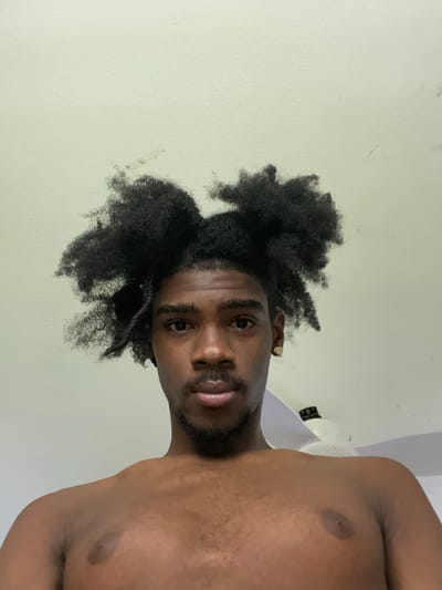 A shirtless man with black, textured hair is looking down at the camera. He has a dark complexion, facial hair around his mouth and on his chin, and an earring in his left ear. The background is a light-colored wall and a ceiling fan. Location: Capesterre-Belle-Eau, France.