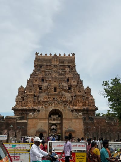 This media file captures a vertical, eye-level shot of the Brihadeeswarar Temple in Thanjavur, India, bathed in daylight under an overcast sky. The main subject is the intricately carved stone tower of the temple, known as the Vimana. Numerous small sculptures of deities, figures, and decorative motifs adorn its facade, showcasing detailed craftsmanship.
To the right of the temple, a crowd of people is visible, suggesting a busy day at the historical site. They appear to be queuing or observing the temple. The people are dressed in various casual attire. Visible text in Tamil is present on yellow signs, some of which are partially obscured by the people. One sign appears to read "அருள்மிகு தஞ்சை தியர் கோவில்" (Arulmigu Thanjai Thevar Kovil), which translates to "Graceful Thanjavur Thevar Temple." Another sign includes "தஞ்சாவூர், இராமநாதபுரம், காரைக்குடி, திருச்சி," listing place names. There's also text that reads "நகிழி இல்லா பகுதி" (Nakizhilaa Paguthi), meaning "Plastic Free Zone."
The overall scene conveys a sense of cultural and historical significance, with a blend of ancient architecture and contemporary visitor activity. The presence of a tree with green foliage at the top left corner provides a natural element against the stone structure and the sky.