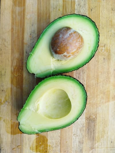 This is a top-down, medium shot of two halves of an avocado placed on a wooden cutting board. The avocado is ripe and its green skin contrasts with its pale yellow flesh. One half of the avocado contains a large, round pit, while the other half is empty, showing the smooth texture of the flesh. The cutting board has a natural wood grain with visible vertical lines and some light staining and scratches. There are a few small water droplets scattered on the cutting board, suggesting it might have been recently used for washing produce. The lighting is even, indicating it's likely an indoor setting, possibly a kitchen. No people are visible in the image. The scene depicts the preparation of food, specifically an avocado, in a domestic or culinary environment. The overall mood is simple, natural, and food-focused. No text is visible in the image.