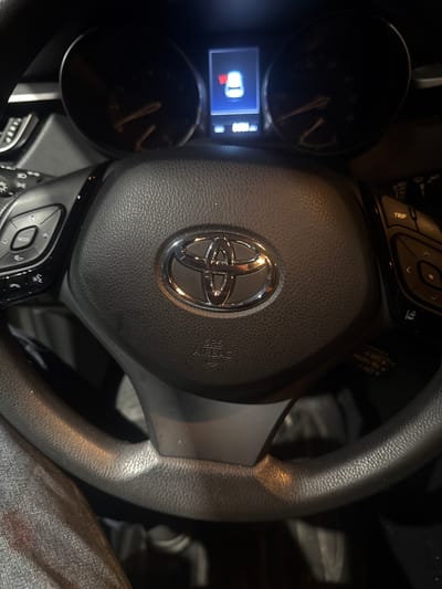 The image shows a close-up of a Toyota steering wheel inside a vehicle. The steering wheel is black leather with a chrome Toyota logo in the center. There are various buttons on the steering wheel, including controls for audio, cruise control, and trip information. In the background, parts of the car's dashboard and instrument cluster are visible. One instrument displays a red icon of a car with an open door. The overall lighting suggests the photo was taken at night or in a dimly lit environment, possibly in Jeffersontown, United States of America, as no other contextual information is provided.