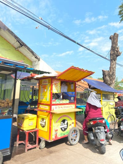 The image shows a street food vendor in Kecamatan Tulangan, Indonesia, a small town located at approximately -7.482 latitude and 112.649 longitude. The vendor is selling iced tea and snacks from a yellow and red cart. The cart is adorned with various Indonesian language signs, including "Es Teh Mu Semaangatku" (Your Iced Tea, Makes You Enthusiastic) and "Segarsnya Bikin Ketagihan" (It's Refreshing, It Makes You Crave More). To the right of the cart is a person seated on a motorcycle, wearing a gray hijab and a maroon long-sleeved shirt, with a blue bag hanging from their left wrist. They are positioned to the right of the cart, near a blue and white, blue and yellow, and a yellow food cart, all equipped with canopies. Behind the carts are two buildings, one with a partially visible bright green wall, and another featuring a yellow roof. The scene is set on a paved road, with electrical wires overhead, and a clear sky with light clouds in the background. A tree is visible in the background, its branches reaching towards the sky.