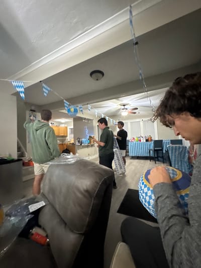 The image depicts an indoor scene, likely a residential living space or common area in South Bend, United States; the exact address within the city cannot be determined from the visual information provided. The environment is decorated with blue and white checkered Bavarian flag-style bunting strung across the ceiling, along with a matching spherical lantern, indicating an Oktoberfest or German-themed event.
Four male individuals are present. In the immediate foreground on the right, a person with dark, curly hair, wearing a grey long-sleeved shirt, is seated or crouched, actively engaged in assembling or inflating a blue and white checkered sphere. To the mid-left, another individual, viewed from behind, wearing a green hooded sweatshirt and light-colored shorts, is standing near a wall, seemingly adjusting or hanging decorations. In the mid-ground center, a third individual, wearing a dark green polo shirt and dark pants, stands holding a clear glass or stein containing a foamy liquid. Adjacent to him and partially obscuring the fourth individual, is a large white promotional bag displaying "Hofbrรคu" text. The fourth individual, in the background right, wears a black t-shirt and light-colored shorts.
The area transitions into a kitchen or bar space in the background, featuring a counter with a sink and blue string lights. Further in the background, a table is covered with a blue and white checkered tablecloth, accompanied by dark chairs. In the left foreground, a grey upholstered recliner is visible, partially covered with clear plastic, on which an orange soda can and a blue packaged item are discernible. The floor is a dark material. The scene conveys a collaborative setup or an early stage of a social gathering.