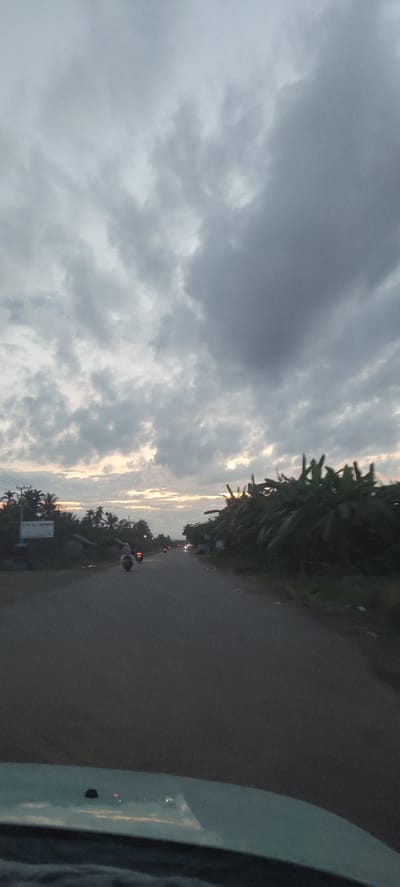 The image depicts a road scene in Meunasahmesjid, Indonesia, likely captured during dusk or early morning. The sky is dominated by heavy, grey clouds, with a subtle strip of warm, orange light visible on the horizon, indicating either sunrise or sunset. The road, which appears to be paved, extends into the distance, with some reflections on its surface in the foreground, suggesting a smooth, possibly wet, texture.
On the left side of the road, various trees and some structures are visible, along with an unidentifiable sign featuring white text on a dark background, the details of which are not legible. A motorcycle with a rider is present in the mid-ground, moving away from the viewer. Further down the road, the faint headlights and taillights of other vehicles suggest ongoing traffic. The right side of the road is lined with dense tropical vegetation, prominently featuring large, lush green leaves resembling those of banana plants. The photograph appears to be taken from inside a vehicle, with a portion of the dashboard or windshield visible in the immediate foreground, showing reflections. The overall ambiance is muted and peaceful, characteristic of twilight hours under an overcast sky.