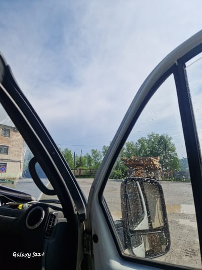 The image is taken from inside a vehicle, looking out through the open driver's side door and window. The sky is blue with scattered clouds. To the left, a brick building with multiple windows is visible. A red and white circular sign with a diagonal red line through it is mounted on a pole in front of the building. In the middle ground, a large yellow mining truck is parked on a dirt road. Behind the truck, a wooden fence runs across the scene, and beyond that, a dense line of green trees. The side mirror of the vehicle is visible in the foreground, reflecting the scene outside, including the yellow truck and the sky. The windows of the vehicle have water droplets on them. The interior of the vehicle shows part of the dashboard and steering wheel. The text "Galaxy S22+" is superimposed in the lower left corner of the image. The overall setting appears to be an industrial or rural area, likely Zheleznogorsk-Ilimsky, Russia, given the context.