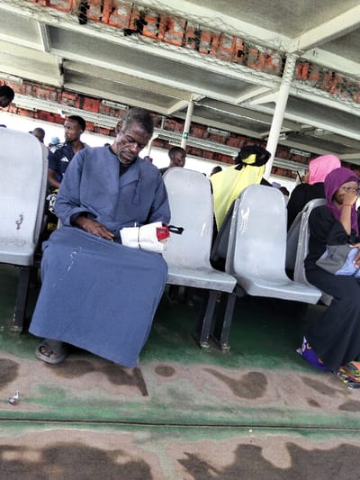 The scene is onboard a passenger ferry or large boat, observed within the Mkomani ward vicinity, Mombasa, Kenya. An elderly man, appearing to be in his 60s-70s, with dark grey hair and beard, is centrally seated in a dark blue long-sleeved garment (thobe or kanzu) and dark sandals, his head bowed, indicating rest or sleep. He holds a small, white bundled item with a red strap. White splatter marks are visible on his garment. To his immediate right, a younger woman in a black garment and purple hijab is seated, looking towards the right of the frame, with a grey bag resting on her lap. Other passengers are visible in the background, including a man in a dark blue polo shirt and individuals wearing pink and yellow head coverings. The environment features rows of light grey plastic seats with dark metal frames, a wet, dark green deck floor with irregular darker stains, and an overhead structure consisting of white beams, support poles, and safety netting containing multiple orange life jackets. A small piece of litter, silver and red, lies on the deck near the central man's feet.