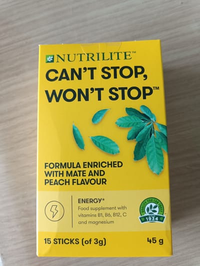 The image displays the front of a bright yellow box of "NUTRLITE CAN'T STOP, WON'T STOP" energy food supplement sticks. The box features prominent black text with the product name. Green leaves are depicted on the front of the box, and there is text indicating it is a formula enriched with mate and peach flavor. A circular emblem with the text "QUALITY YOU CAN TRUST SINCE 1934" is also visible. Below the main text, it states "ENERGY*" and lists that it is a food supplement with vitamins B1, B6, B12, C, and magnesium. The bottom of the box indicates "15 STICKS (of 3g)" and "45 g". The box is placed on a light-colored wooden surface. There are no people or animals present in the image, and no context related to Sant'Andrea in Besanigo, Italy is provided or inferable from the image itself.
