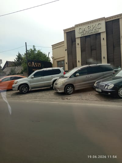 The image depicts a scene in front of a building with a sign reading "OASIS РЕСТОРАНЫ" in Cyrillic, and "OASIS" in English. The building appears to be a restaurant or entertainment venue, with a beige facade and dark vertical panels. In front of the building, several cars are parked on a gravel lot. These include a silver SUV, a silver minivan, and a dark sedan. To the left of these vehicles, an orange car is partially visible. In the background, there are trees and residential buildings, suggesting a mixed-use area. The sky is overcast, and the overall lighting is dim. The timestamp "19.06.2026 11:54" is visible in the lower right corner. The location of Romanovka, Kyrgyzstan is not explicitly indicated in the image itself, but is provided as context. There are no people visible in the frame.