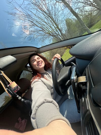 A young woman with reddish-brown hair and fair skin is taking a selfie inside a car. She is in the driver's seat, holding the steering wheel and smiling at the camera. She is wearing a gray hooded sweatshirt and blue jeans. The car's interior is visible, including the dashboard and windshield. Outside, trees and a blue sky are seen through the windshield, potentially indicative of an outdoor setting. There is no definitive information to confirm the location as the city of Cheryomushki, Russia. Other objects, such as a car phone holder, are visible as well as what appears to be brushes or decorative items.